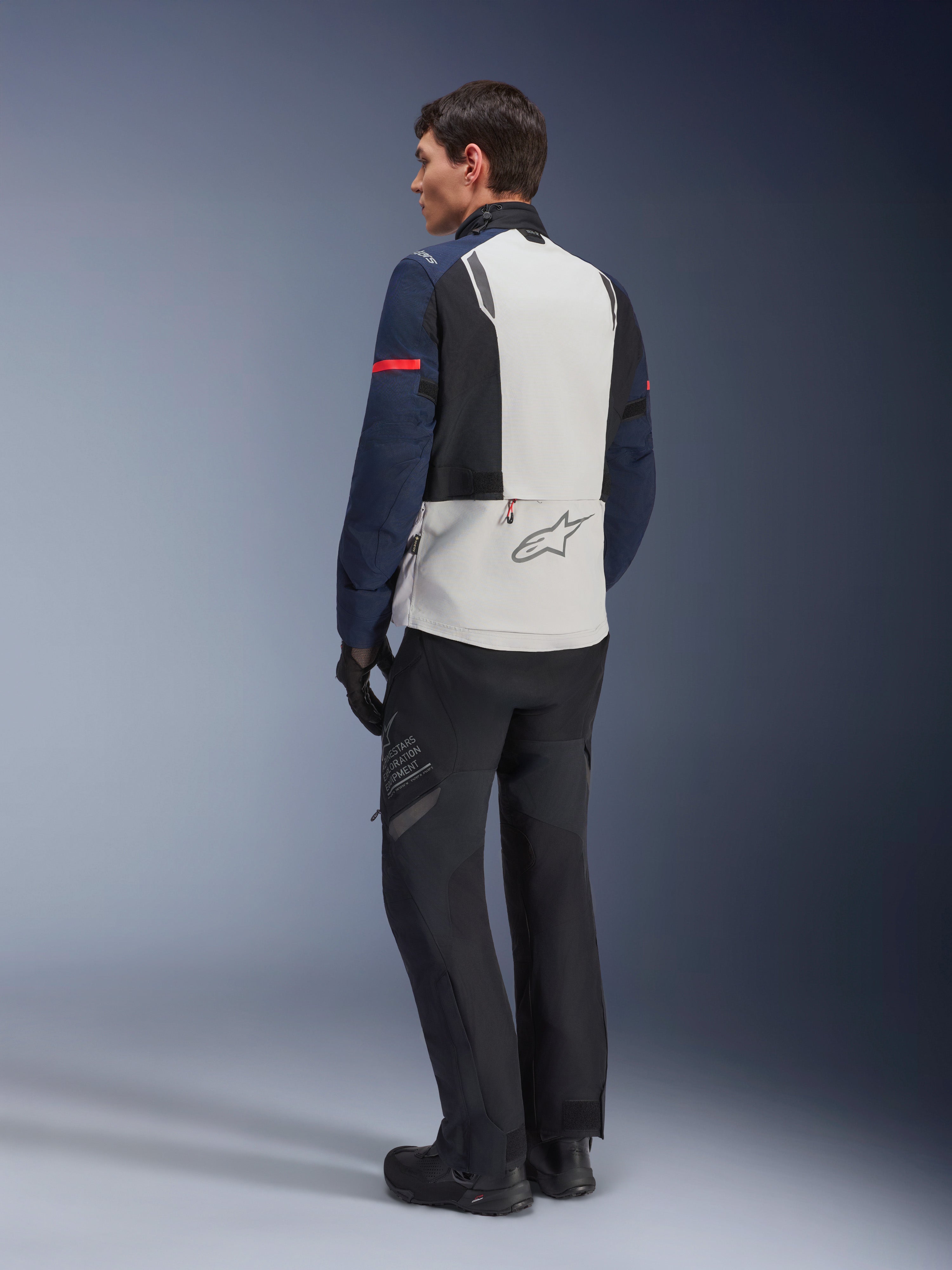 Modello che indossa la giacca Alpinestars ST-7 2L GORE-TEX in Ice Gray Dark Blue e Black, vista posteriore, pannello posteriore grigio chiaro con logo Alpinestars, maniche blu scuro con striscia rossa, abbinata a pantaloni e stivali da moto adventure neri, abbigliamento da moto touring