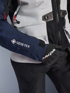 Giacca Alpinestars ST-7 2L GORE-TEX, primo piano della manica blu scuro con logo GORE-TEX bianco, busto grigio ghiaccio con dettagli neri e accenti in cordino rosso, indossata con guanti e pantaloni neri Alpinestars, abbigliamento da moto adventure touring