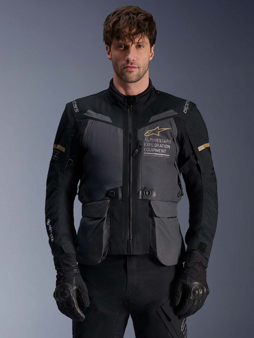 Giacca Alpinestars ST-7 2L GORE-TEX, giacca da moto adventure touring da uomo, grigio scuro e nero con dettagli kaki, con marchio 'Alpinestars Exploration Equipment', tasche cargo impermeabili e collo alto, indossata da un modello con guanti neri