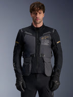 Giacca Alpinestars ST-7 2L GORE-TEX, giacca da moto adventure touring da uomo, grigio scuro e nero con dettagli kaki, con marchio 'Alpinestars Exploration Equipment', tasche cargo impermeabili e collo alto, indossata da un modello con guanti neri