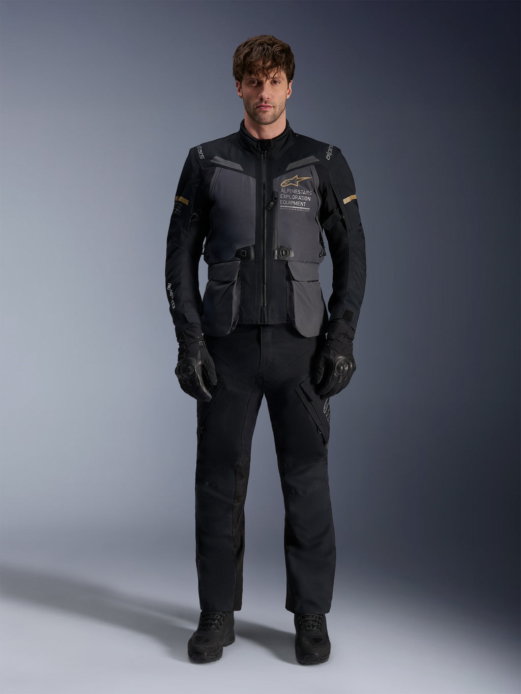 Modello che indossa la giacca Alpinestars ST-7 2L GORE-TEX in Deep Gray Black Dark Khaki, con il marchio 'ALPINESTARS EXPLORATION EQUIPMENT' sul petto, abbinata a pantaloni da moto, guanti e stivali neri coordinati, vista frontale in studio.