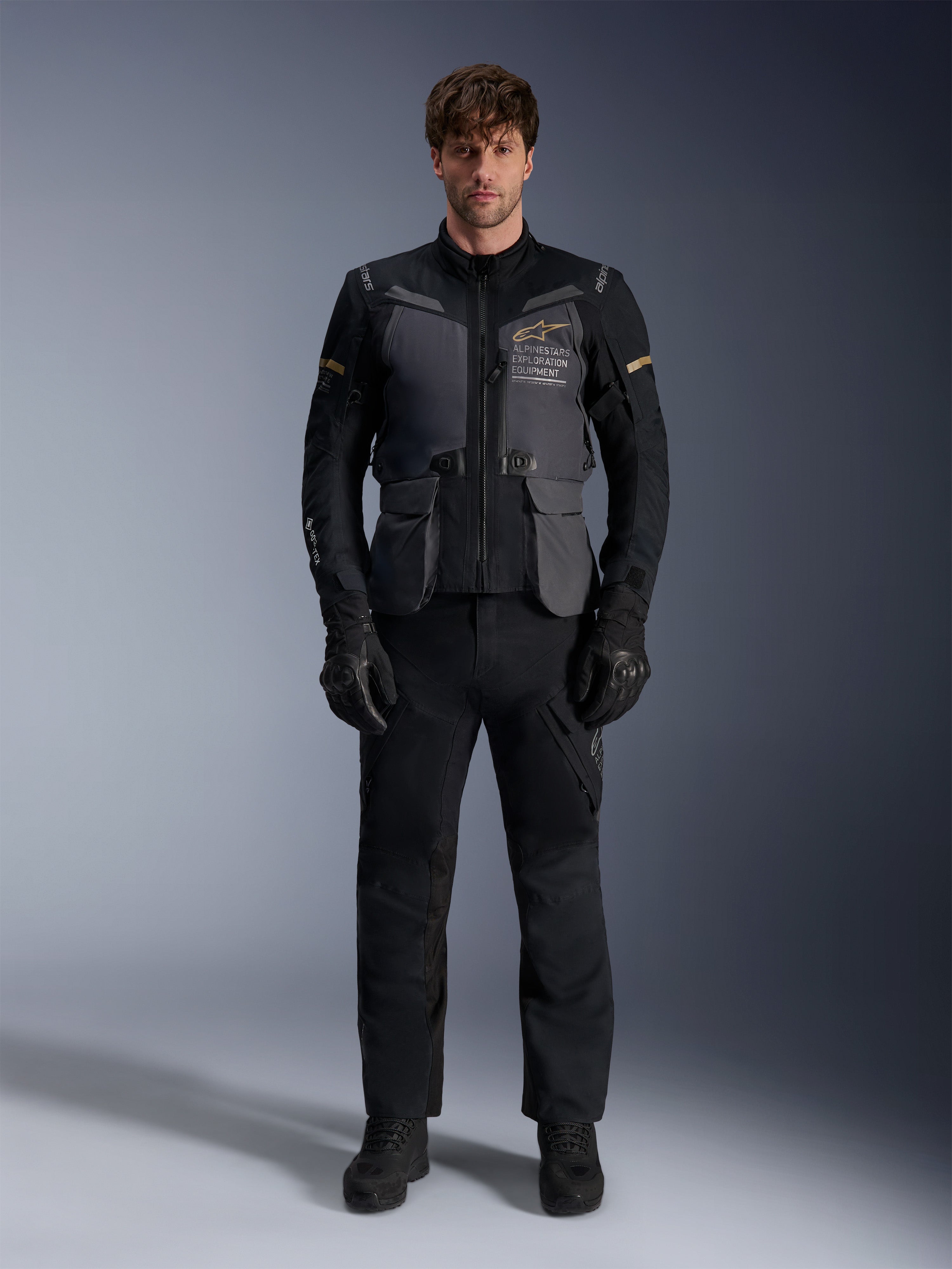 Modello che indossa la giacca Alpinestars ST-7 2L GORE-TEX in Deep Gray Black Dark Khaki, con il marchio 'ALPINESTARS EXPLORATION EQUIPMENT' sul petto, abbinata a pantaloni da moto, guanti e stivali neri coordinati, vista frontale in studio.