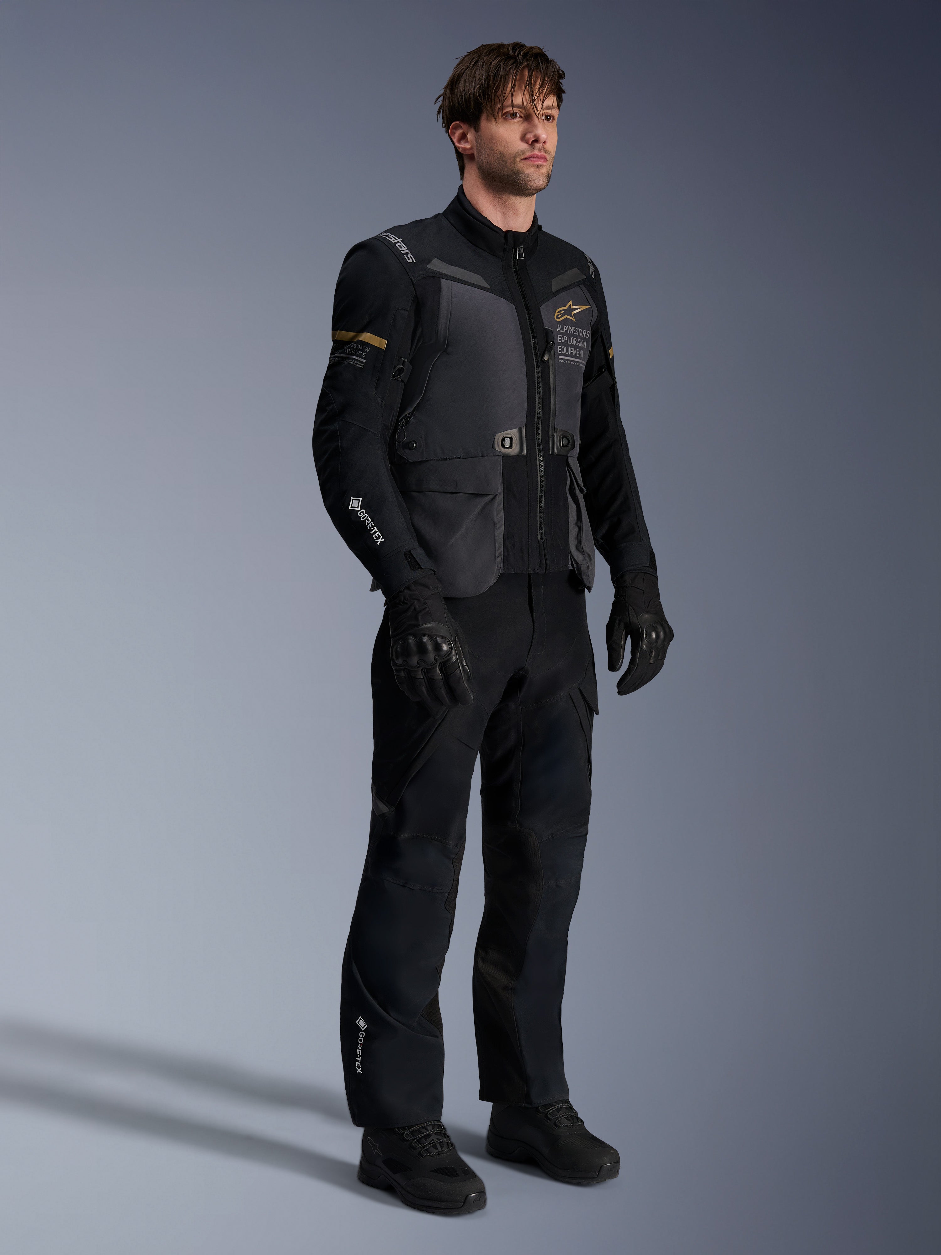 Modello che indossa la giacca Alpinestars ST-7 2L GORE-TEX in Deep Gray Black e Dark Khaki, abbinata a pantaloni e guanti adventure neri coordinati, con guscio laminato impermeabile, tasche cargo e branding tecnico, ideale per l'adventure touring.