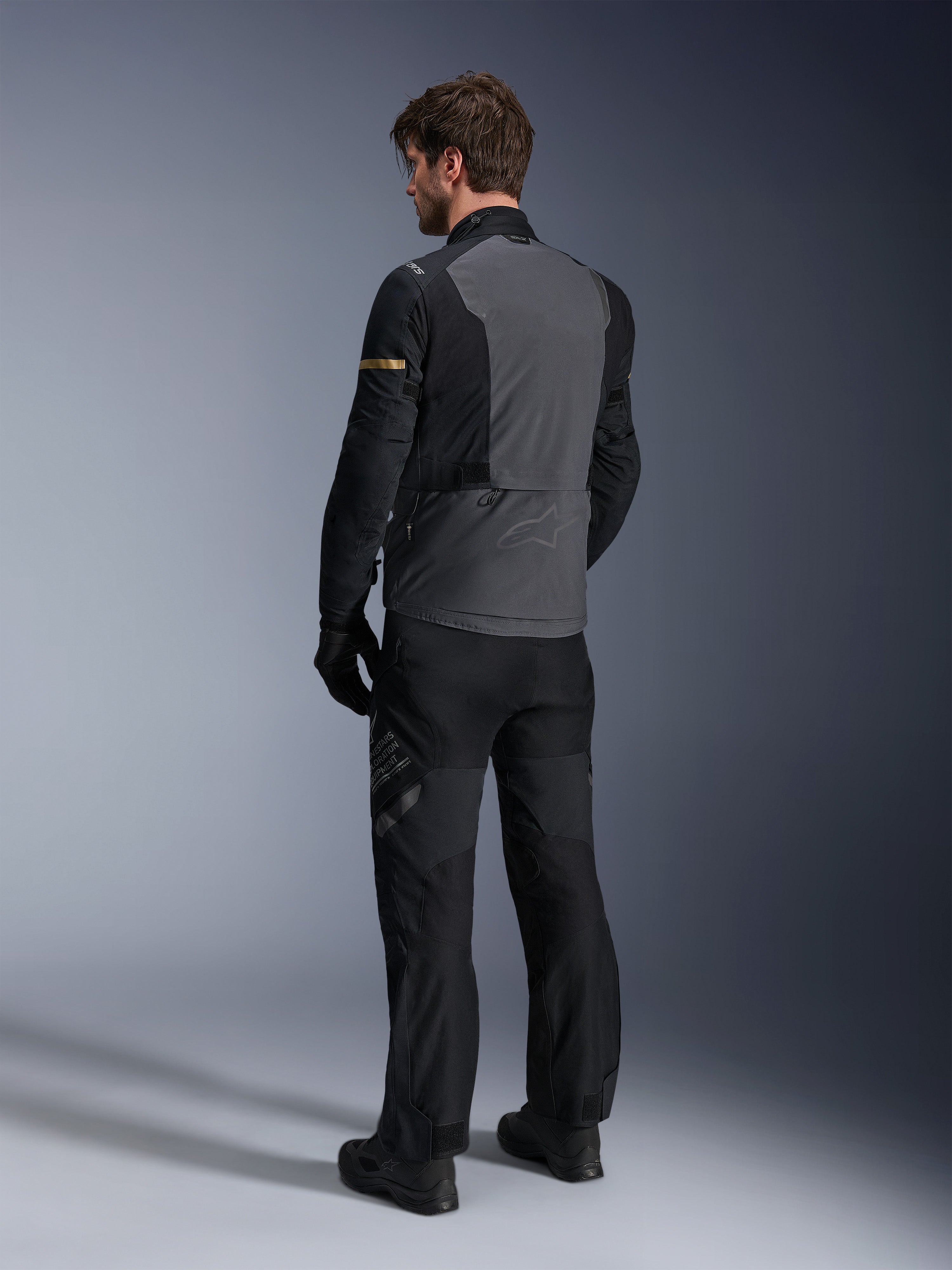 Giacca Alpinestars ST-7 2L GORE-TEX, Deep Gray e nero con accenti Dark Khaki, vista posteriore di un pilota che indossa la giacca e i pantaloni da moto coordinati, abbigliamento adventure touring con costruzione laminata impermeabile