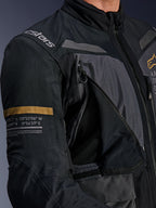 Giacca Alpinestars ST-7 2L GORE-TEX, grigio scuro e nero, primo piano con pannello di ventilazione sul petto aperto con zip e mesh, logo Alpinestars sulla spalla e striscia khaki sulla manica con coordinate geografiche stampate, abbigliamento tecnico da moto adventure-touring