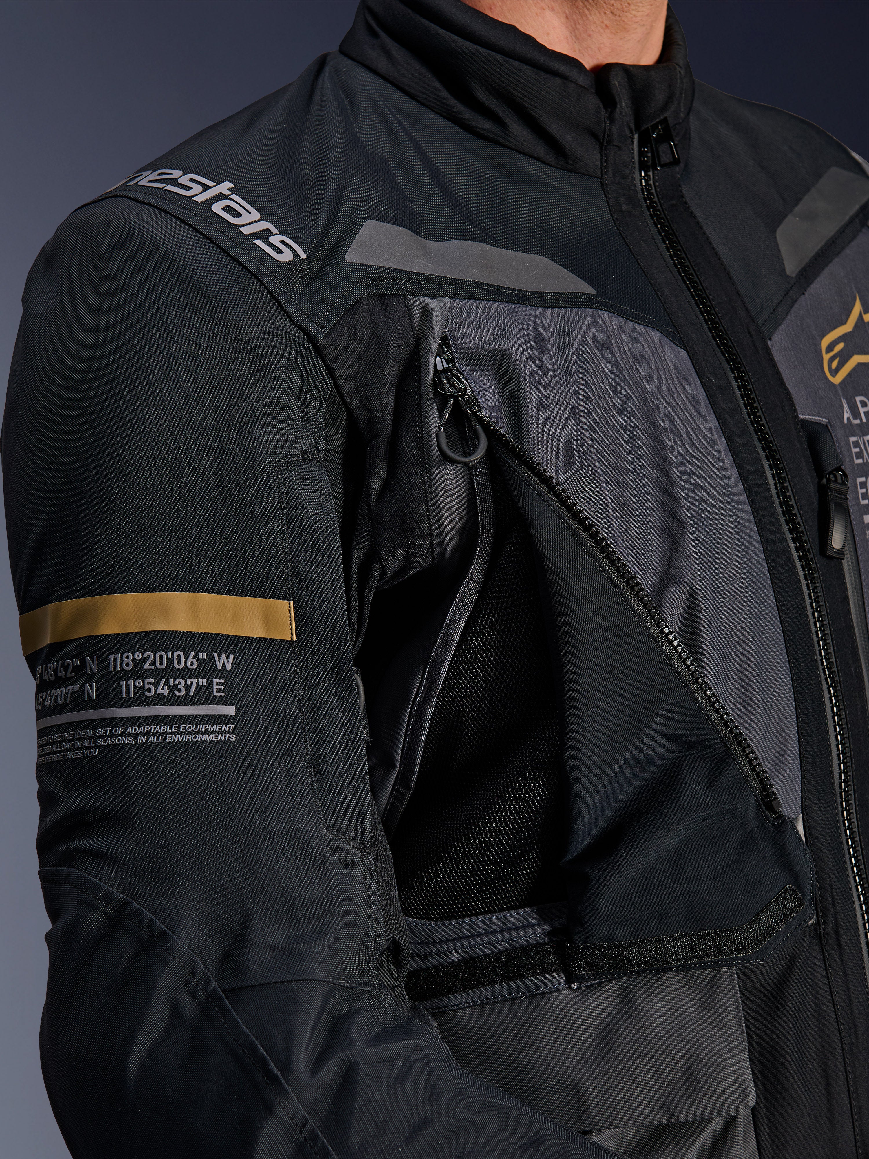 Giacca Alpinestars ST-7 2L GORE-TEX, grigio scuro e nero, primo piano con pannello di ventilazione sul petto aperto con zip e mesh, logo Alpinestars sulla spalla e striscia khaki sulla manica con coordinate geografiche stampate, abbigliamento tecnico da moto adventure-touring