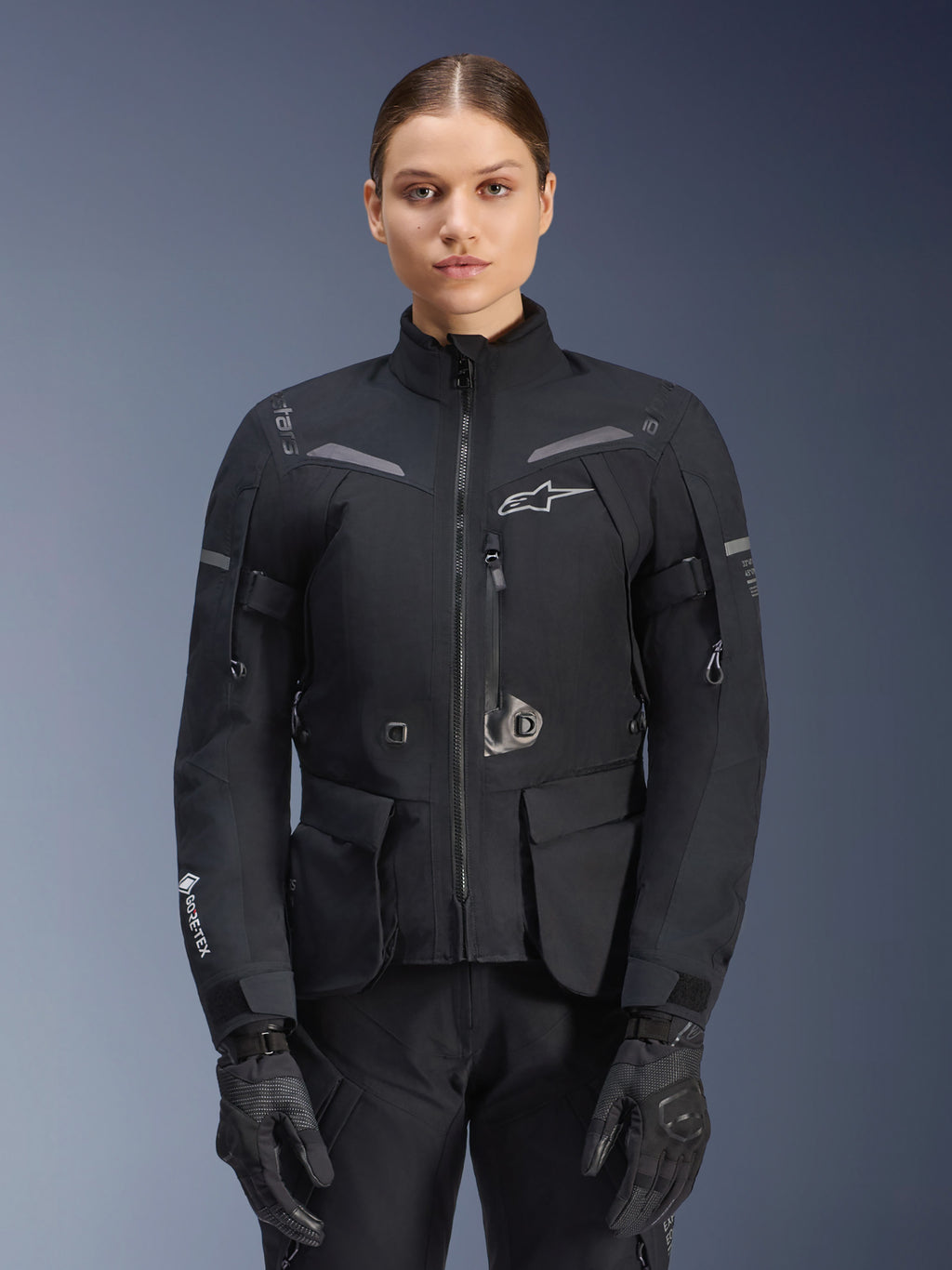 Donna che indossa la giacca Alpinestars Stella ST-7 2L GORE-TEX, nera con dettagli grigio scuro, giacca da moto touring con ampie tasche cargo e collo alto, abbinata a guanti e pantaloni neri, abbigliamento impermeabile per guida adventure