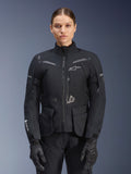 Donna che indossa la giacca Alpinestars Stella ST-7 2L GORE-TEX, nera con dettagli grigio scuro, giacca da moto touring con ampie tasche cargo e collo alto, abbinata a guanti e pantaloni neri, abbigliamento impermeabile per guida adventure