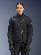 Donna che indossa la giacca Alpinestars Stella ST-7 2L GORE-TEX, nera con dettagli grigio scuro, giacca da moto touring con ampie tasche cargo e collo alto, abbinata a guanti e pantaloni neri, abbigliamento impermeabile per guida adventure