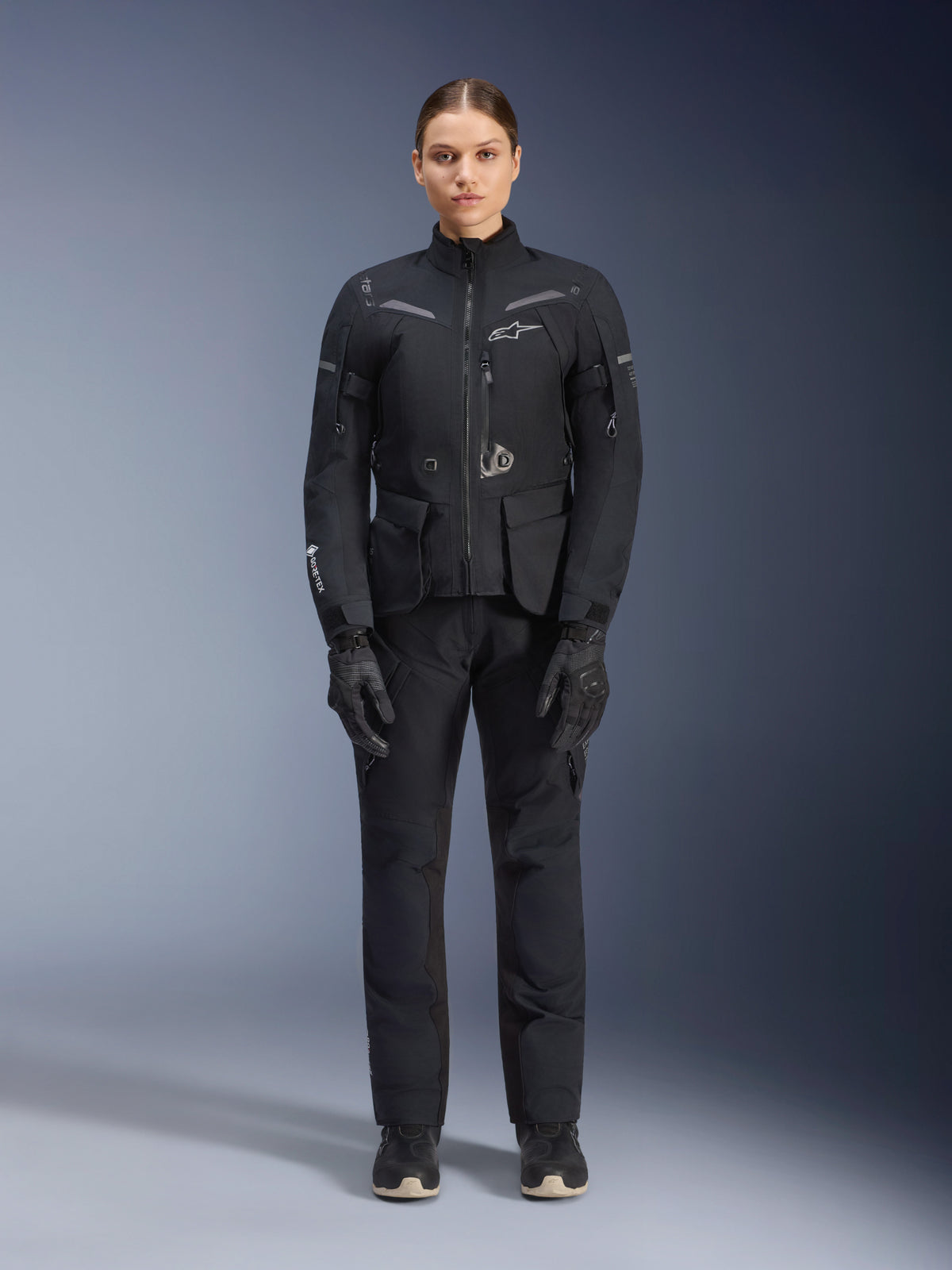 Motociclista donna con giacca e pantaloni Alpinestars Stella ST-7 2L GORE-TEX, completo da touring nero, abbigliamento tecnico impermeabile e traspirante, indossato con guanti e stivali neri coordinati, protezione completa con elementi riflettenti, design tecnico moderno per l'adventure riding