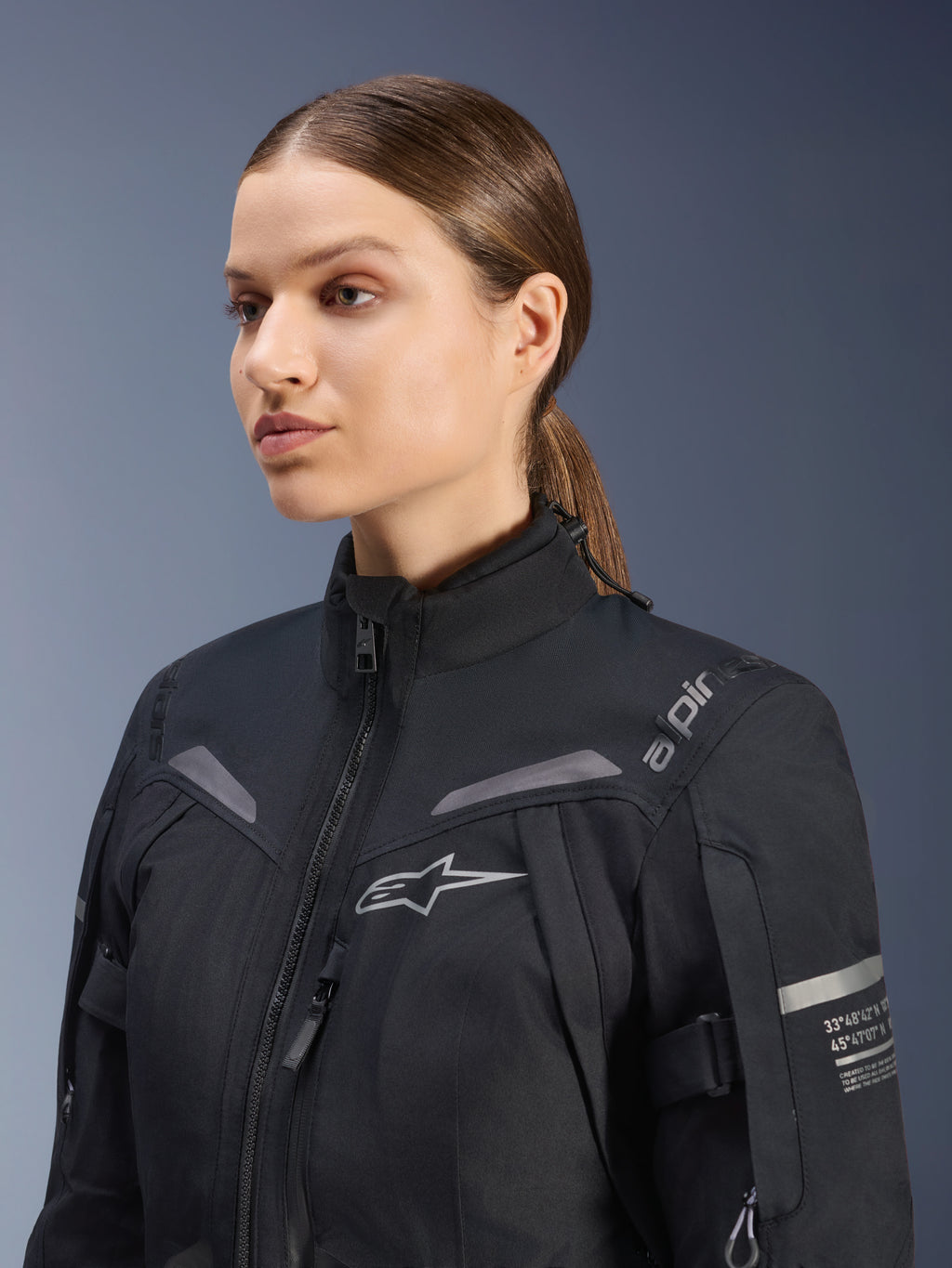 Giacca Alpinestars Stella ST-7 2L GORE-TEX da donna, nera, con logo sul petto e coordinate tecniche sul braccio, design impermeabile in tessuto a collo alto per mototurismo, ritratto in studio di una motociclista.
