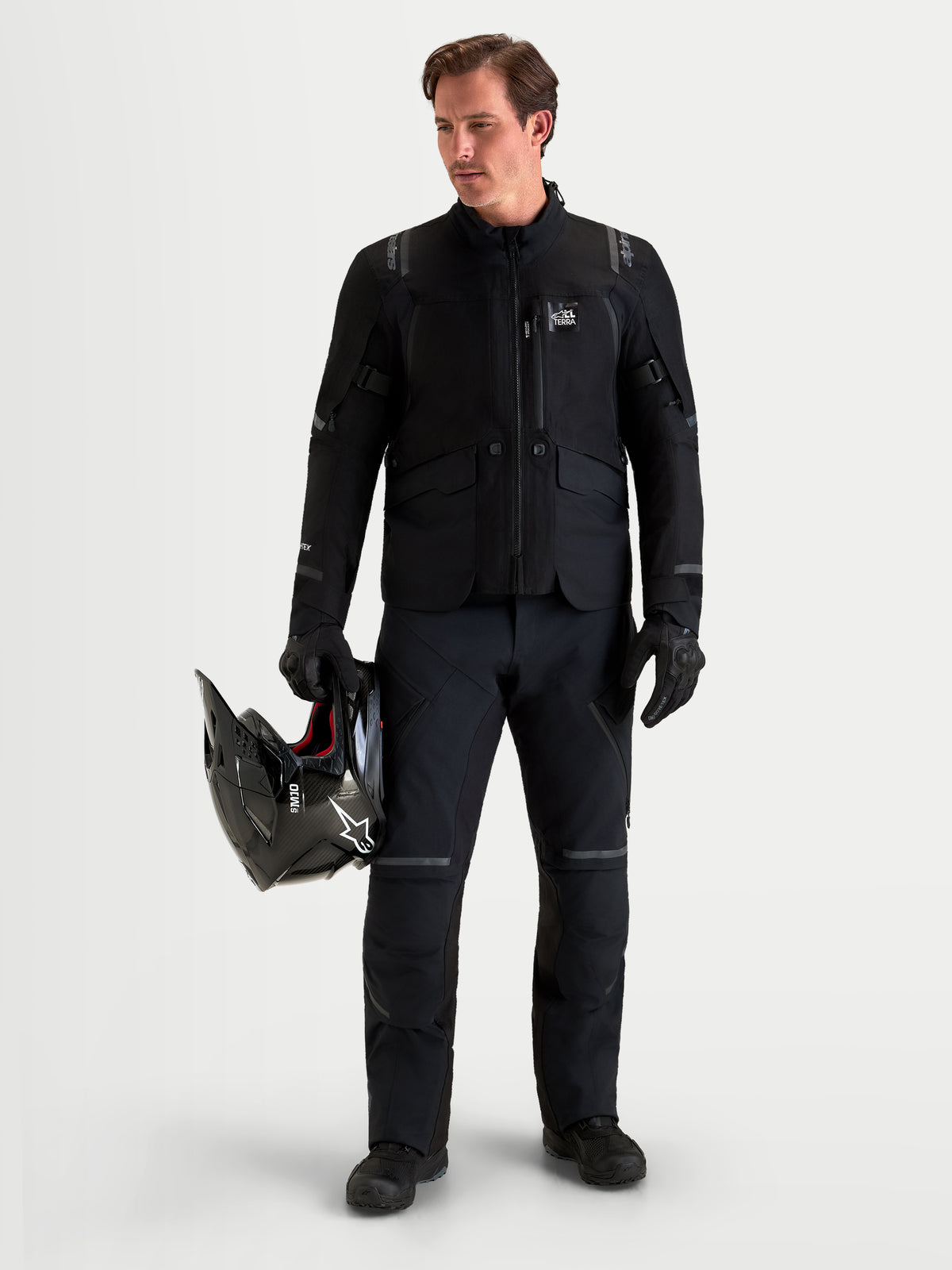 Alpinestars St-7 V2 2L Gore-Tex Pantaloni Short, pantaloni da moto adventure da uomo in colore nero, vista frontale su un modello che indossa abbigliamento coordinato e tiene in mano un casco, abbigliamento di guida all-terrain impermeabile certificato AA