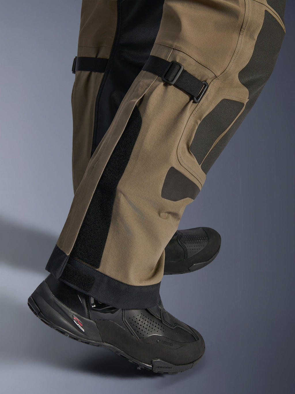 Primo piano dei pantaloni Alpinestars Nazca 3L GORE-TEX PRO in Walnut Black, con dettagli tecnici della parte inferiore della gamba inclusi i regolatori di volume del polpaccio e i rinforzi in Superfabric, indossati con stivali da moto adventure neri.