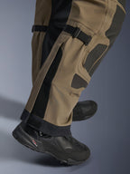 Primo piano dei pantaloni Alpinestars Nazca 3L GORE-TEX PRO in Walnut Black, con dettagli tecnici della parte inferiore della gamba inclusi i regolatori di volume del polpaccio e i rinforzi in Superfabric, indossati con stivali da moto adventure neri.