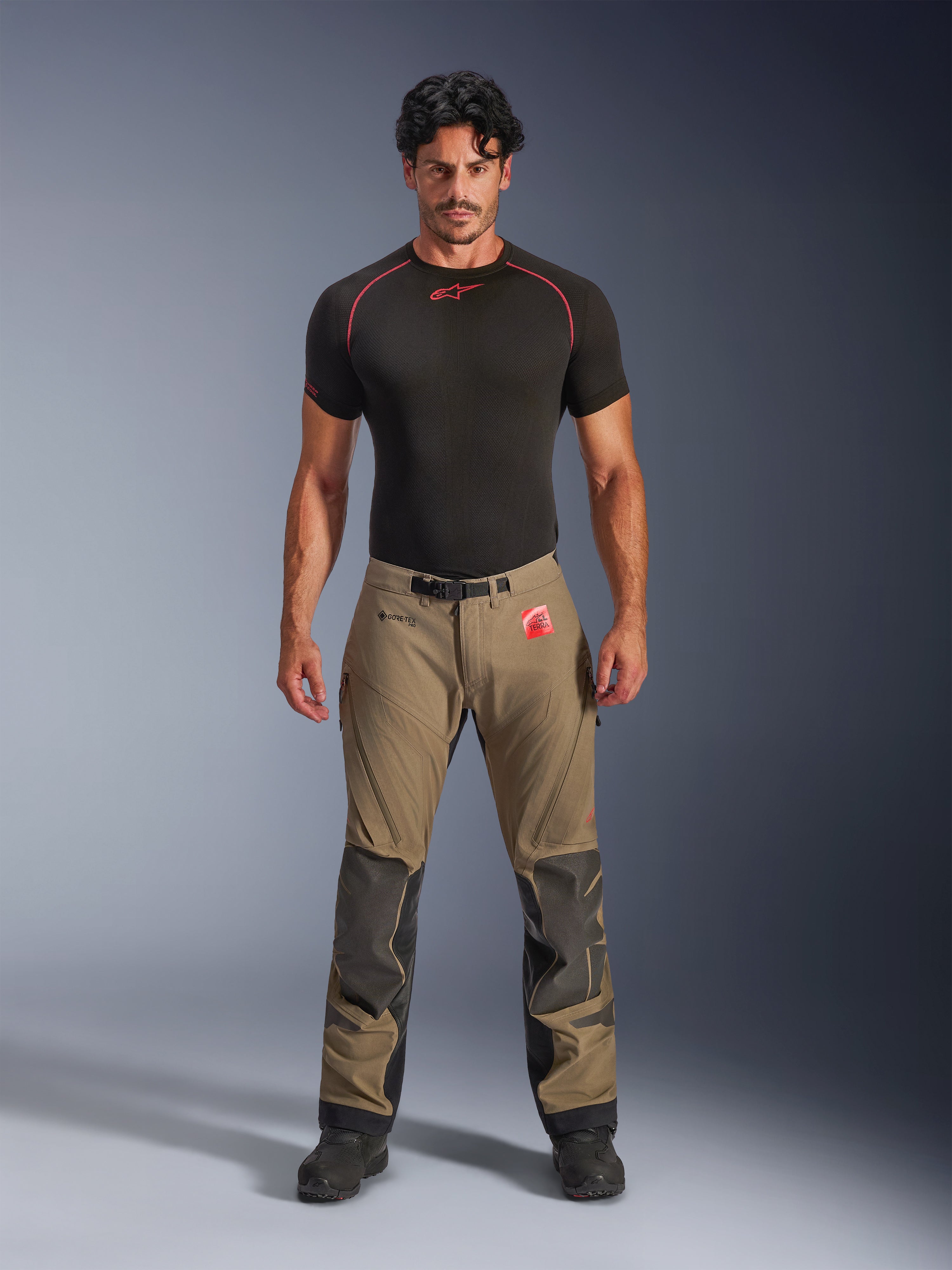 Modello che indossa pantaloni Alpinestars Nazca 3L GORE-TEX PRO in marrone noce e nero, abbigliamento tecnico da moto adventure con rinforzi sulle ginocchia in Superfabric e fibbia magnetica in vita, abbinati a una maglia base layer Alpinestars nera, vista frontale in studio.