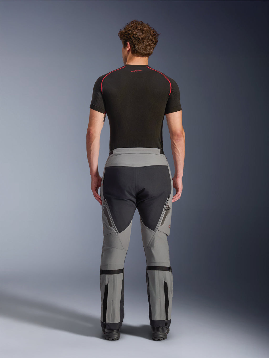Pantaloni Alpinestars Nazca 3L GORE-TEX PRO, pantaloni da moto adventure in grigio scuro e nero, vista posteriore su un modello, design tecnico multi-pannello con tessuto grigio e nero, con regolatori di volume del polpaccio e indossati con uno strato base nero con accenti rossi.