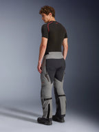 Pantaloni Alpinestars Nazca 3L GORE-TEX PRO, pantaloni da adventure riding, grigio scuro e nero, vista posteriore di un modello che indossa un base layer nero e stivali, con dettagli su pannelli tecnici, cerniere di ventilazione e regolatori di volume del polpaccio.