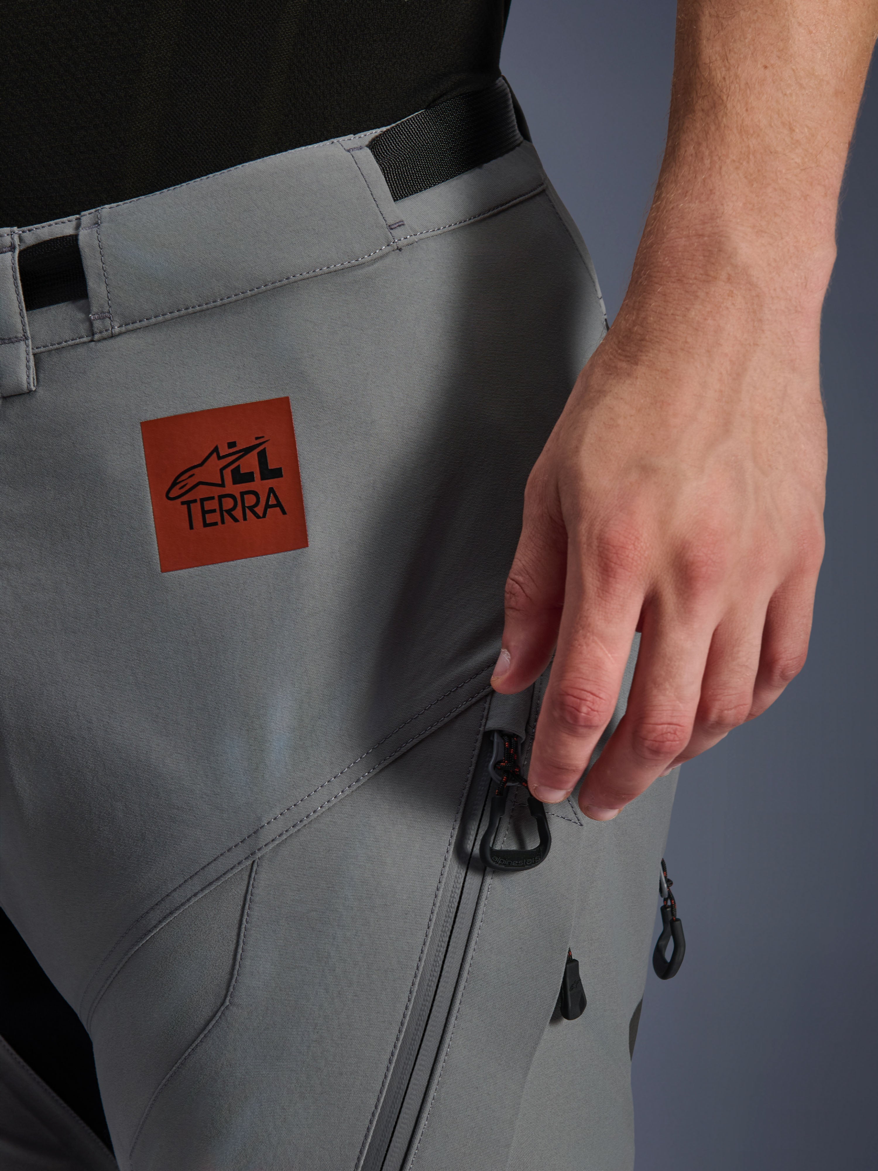 Pantaloni Alpinestars Nazca 3L GORE-TEX PRO, pantaloni da moto adventure, grigio scuro, primo piano della parte superiore della coscia con toppa logo All Terra marrone rossastro, tasca cargo con zip e tiretti neri, robusta costruzione in tessuto per la guida su ogni terreno