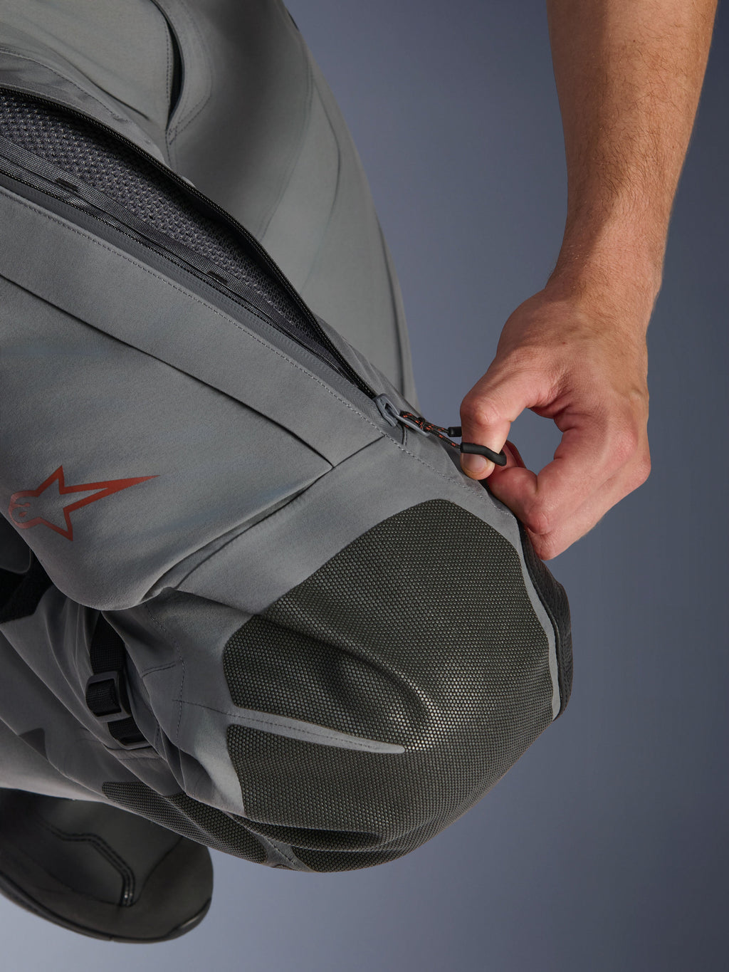 Alpinestars Nazca 3L GORE-TEX PRO Pants, pantaloni da moto adventure, grigio scuro e nero, primo piano di una mano che apre la presa d'aria con zip sulla coscia, con rinforzi per le ginocchia in Superfabric testurizzato e logo a stella Alpinestars rosso, costruzione in tessuto tecnico per protezione in ogni condizione atmosferica