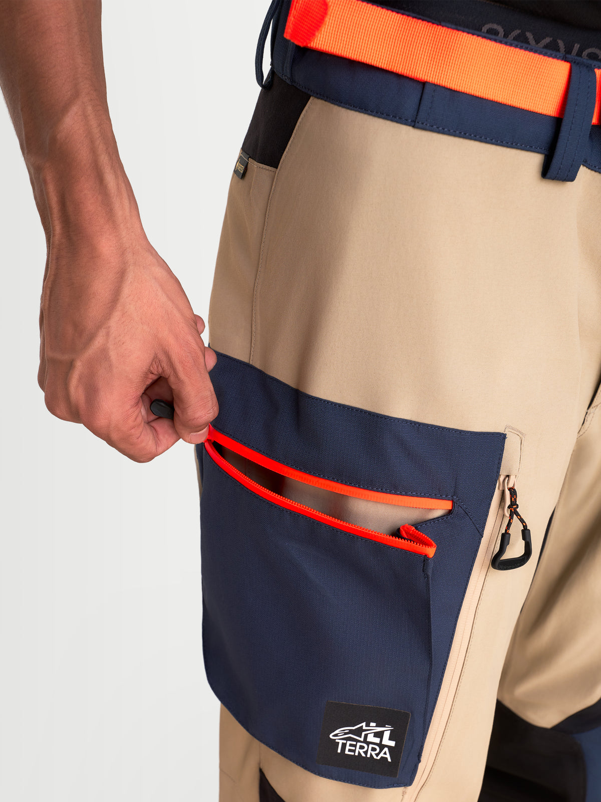 Alpinestars Arenis Gore-Tex Pantaloni, pantaloni da moto adventure, vista ravvicinata con una mano che apre la tasca cargo integrata con dettagli arancioni brillanti, struttura in tessuto impermeabile beige e blu scuro