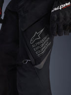 Pantaloni Alpinestars ST-7 2L GORE-TEX in nero, vista dettagliata della tasca cargo sulla coscia con marchio grigio Alpinestars Exploration Equipment, logo a stella e coordinate geografiche, costruzione in tessuto tecnico per adventure touring