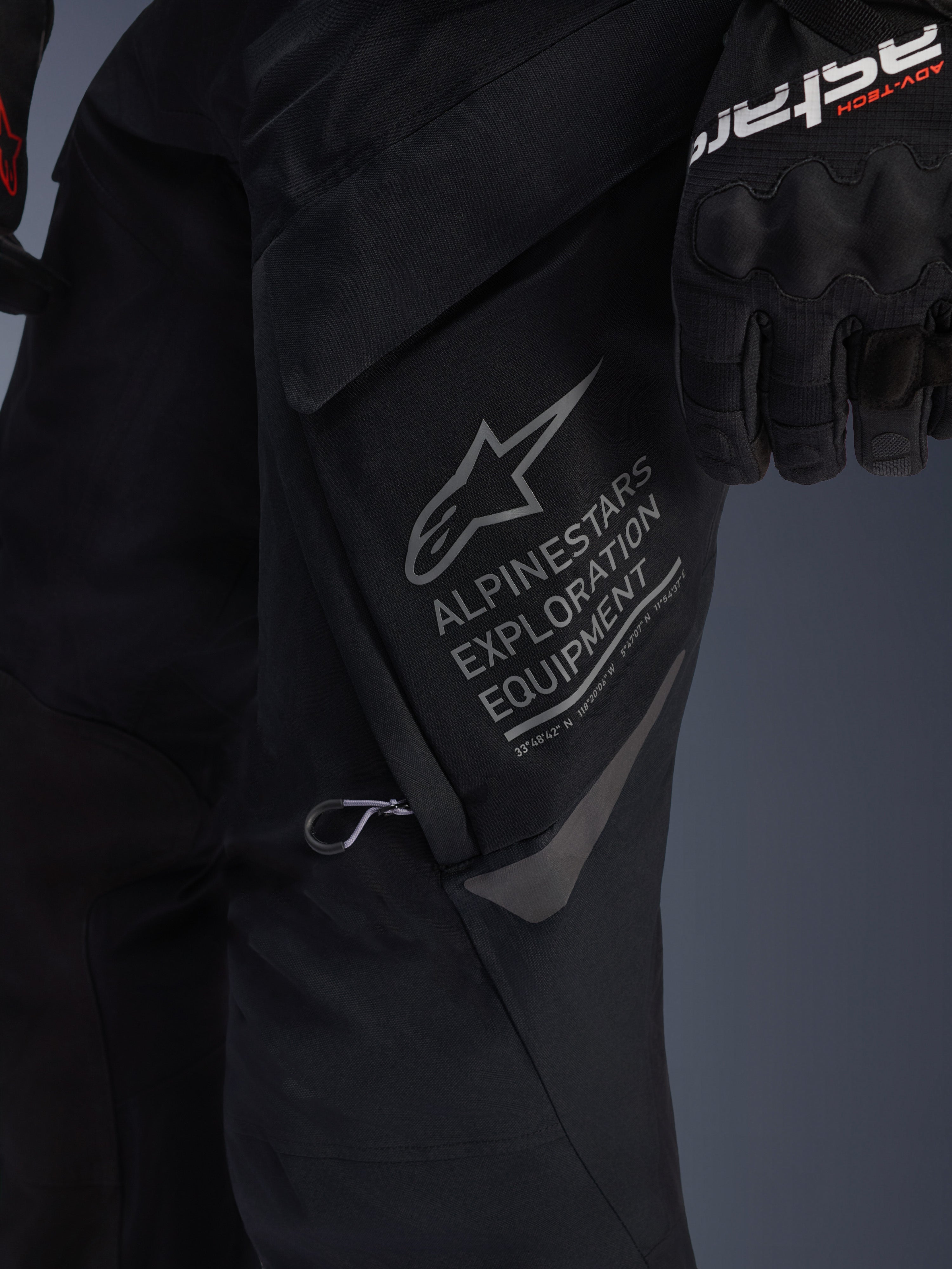 Pantaloni Alpinestars ST-7 2L GORE-TEX in nero, vista dettagliata della tasca cargo sulla coscia con marchio grigio Alpinestars Exploration Equipment, logo a stella e coordinate geografiche, costruzione in tessuto tecnico per adventure touring
