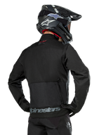 Vista posteriore di un pilota che indossa la giacca Alpinestars Pro-Dura in nero, giacca da enduro in tessuto ripstop resistente, caratterizzata da una grande tasca cargo posteriore con marchio Alpinestars grigio sulla parte bassa della schiena, abbinata a un casco da motocross nero.