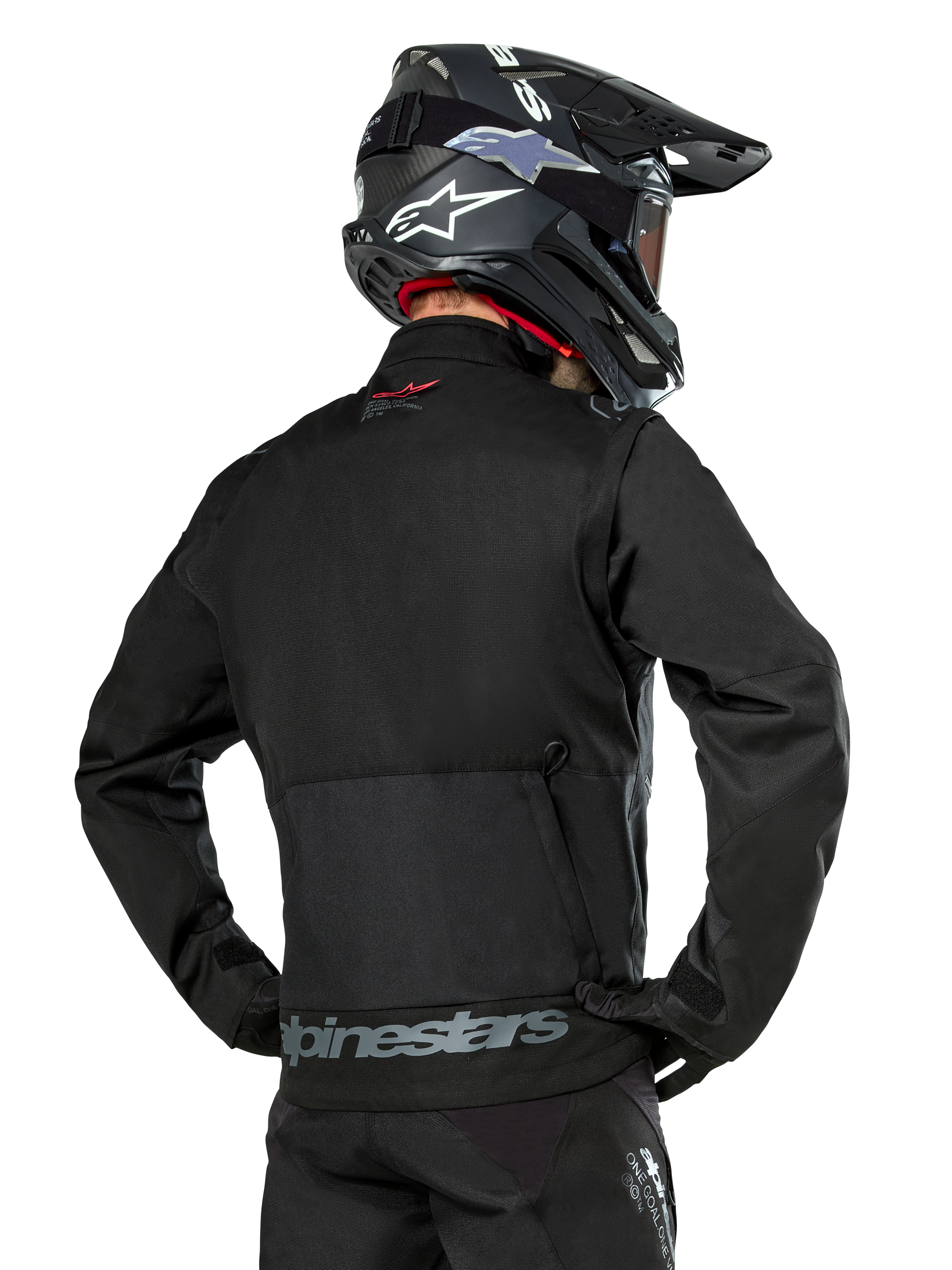 Vista posteriore di un pilota che indossa la giacca Alpinestars Pro-Dura in nero, giacca da enduro in tessuto ripstop resistente, caratterizzata da una grande tasca cargo posteriore con marchio Alpinestars grigio sulla parte bassa della schiena, abbinata a un casco da motocross nero.