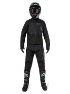 Giacca Alpinestars Pro-Dura, giacca nera da enduro off-road, mostrata su un modello con un set completo di attrezzatura che include casco nero con maschera rossa, pantaloni neri con dettagli rossi, stivali neri e bianchi e guanti neri, design robusto ispirato al rally per trail riding