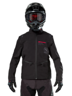 Pilota di motocross che indossa la Giacca Softshell Alpinestars Lite-Dura in nero, con dettagli del logo rossi e grigi sul petto, casco nero con occhiali dalle lenti rosse, guanti e pantaloni neri, vista frontale di abbigliamento tecnico da fuoristrada
