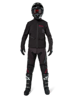 Pilota di motocross con Giacca Softshell Alpinestars Lite-Dura in nero e dettagli del logo rossi, abbinata a pantaloni off-road neri coordinati, casco nero e stivali bianchi e neri, abbigliamento professionale completo da enduro