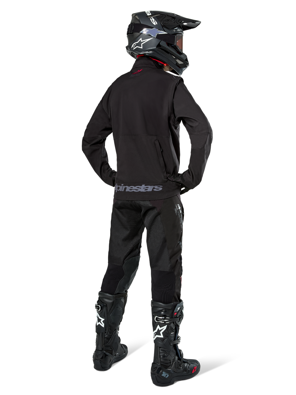 Pilota di enduro visto di schiena con la giacca Softshell Alpinestars Lite-Dura in nero, abbinata a pantaloni tecnici e stivali neri, con tasca posteriore con zip e logo Alpinestars sull'orlo inferiore, equipaggiamento completo off-road per motocross.