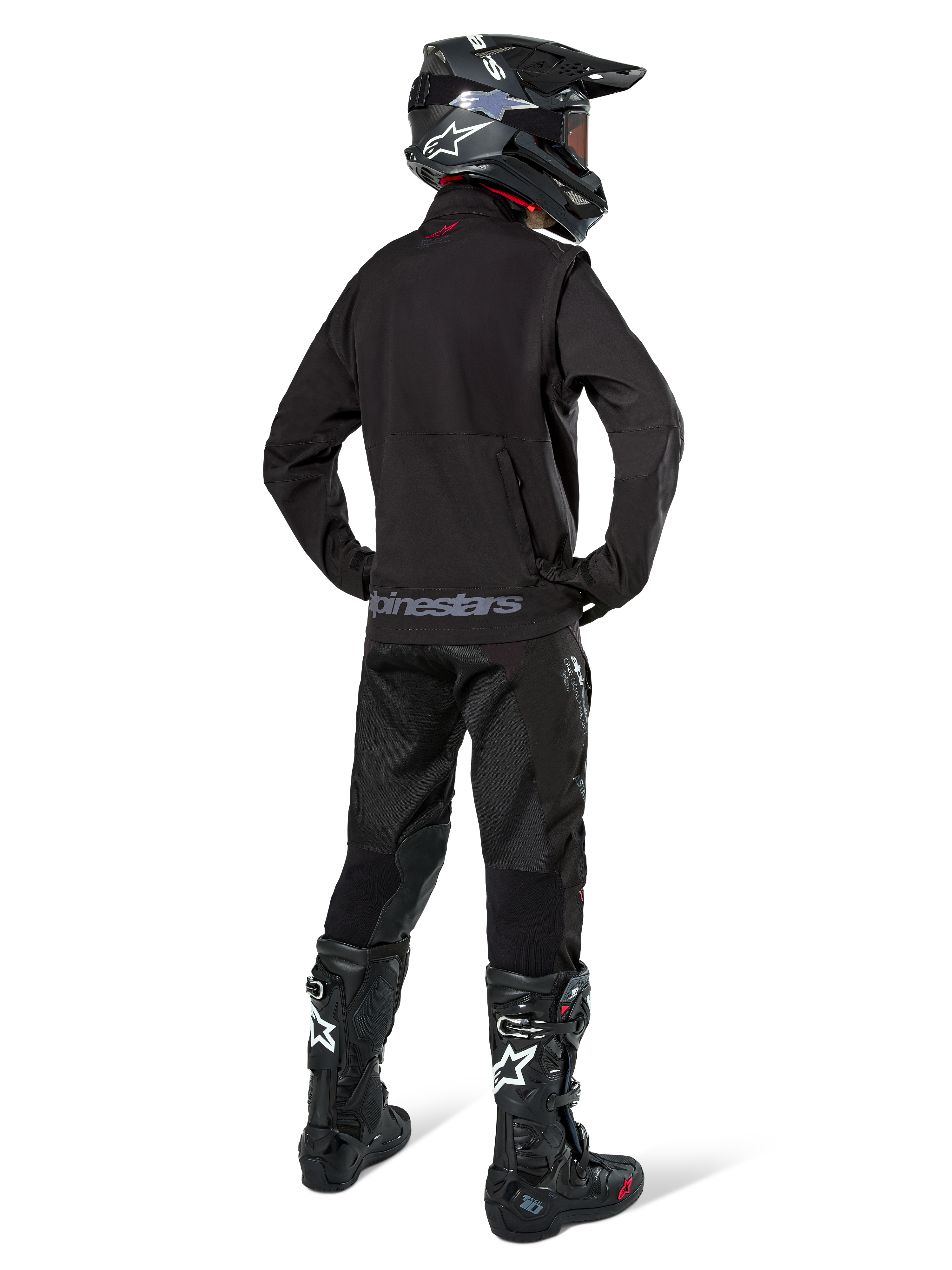 Pilota di enduro visto di schiena con la giacca Softshell Alpinestars Lite-Dura in nero, abbinata a pantaloni tecnici e stivali neri, con tasca posteriore con zip e logo Alpinestars sull'orlo inferiore, equipaggiamento completo off-road per motocross.