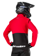 Alpinestars Lite-Dura Softshell Jacket, nero, rosso brillante e bianco, vista posteriore su un pilota di motocross, parte superiore e maniche rosse con pannello posteriore inferiore nero, tasca posteriore portaoggetti con zip, logo Alpinestars bianco sull'orlo inferiore, indossata con casco e pantaloni neri.