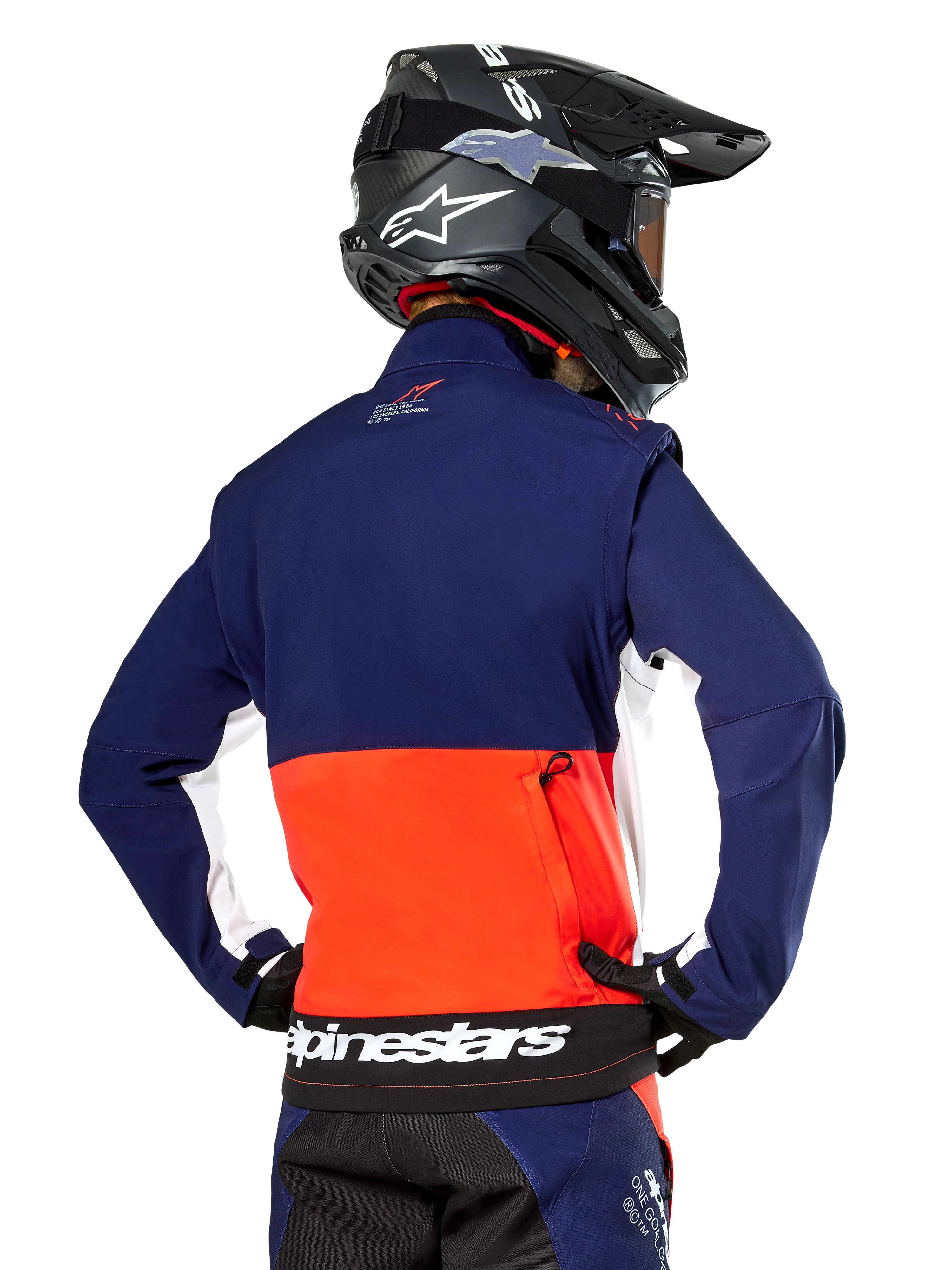 Giacca Softshell Alpinestars Lite-Dura, blu navy, arancione acceso e bianco, vista posteriore indossata da un pilota di motocross, con un grande logo bianco sull'orlo posteriore e tasca portaoggetti con zip, parte di un completo da gara off-road