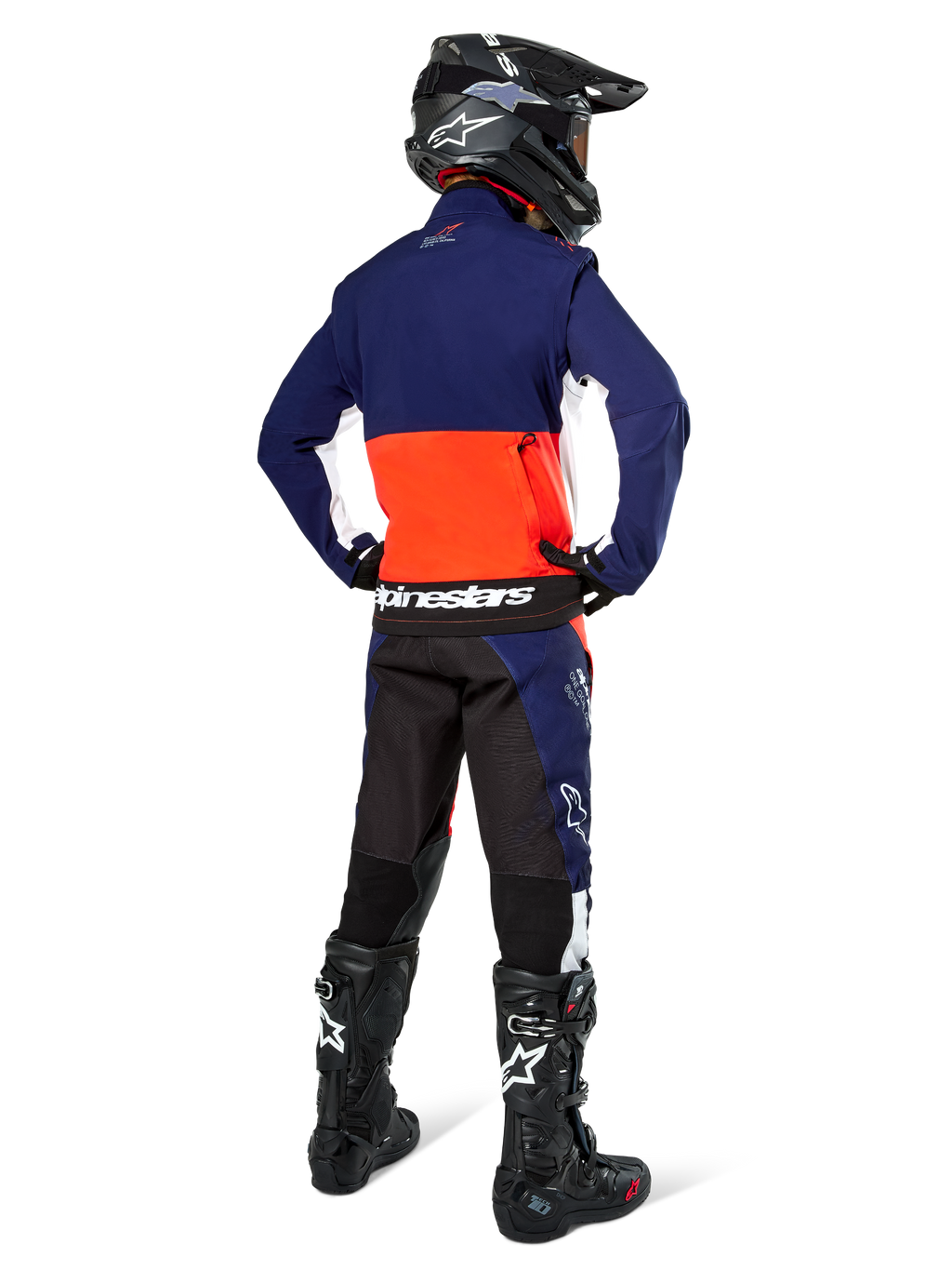 Pilota di motocross visto di spalle che indossa la giacca Alpinestars Lite-Dura Softshell nei colori blu navy, arancione acceso e bianco, mostrando la capiente tasca posteriore. Abbinata a pantaloni da enduro coordinati, casco da motocross nero e stivali da fuoristrada neri.
