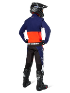 Pilota di motocross visto di spalle che indossa la giacca Alpinestars Lite-Dura Softshell nei colori blu navy, arancione acceso e bianco, mostrando la capiente tasca posteriore. Abbinata a pantaloni da enduro coordinati, casco da motocross nero e stivali da fuoristrada neri.
