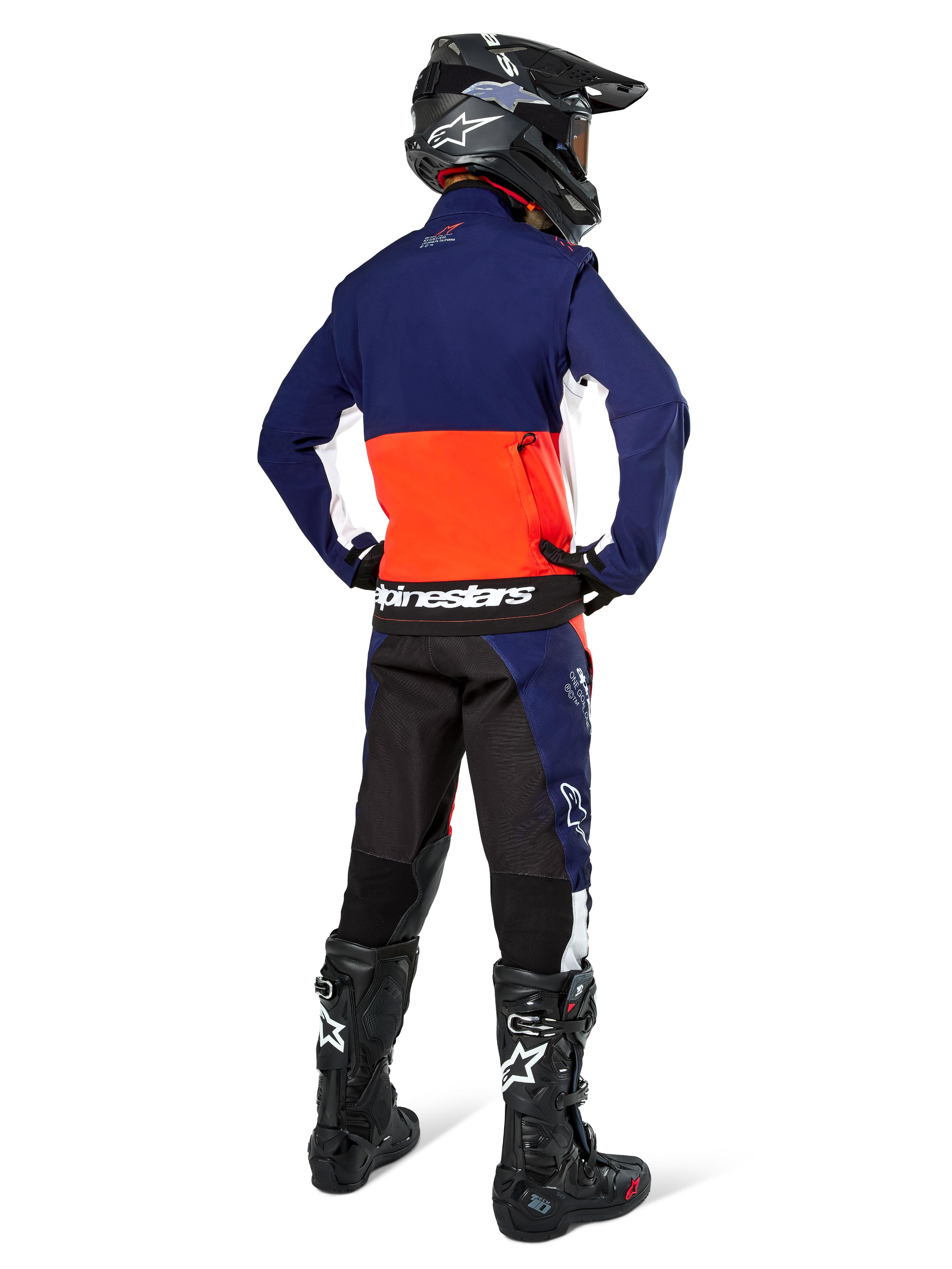 Pilota di motocross visto di spalle che indossa la giacca Alpinestars Lite-Dura Softshell nei colori blu navy, arancione acceso e bianco, mostrando la capiente tasca posteriore. Abbinata a pantaloni da enduro coordinati, casco da motocross nero e stivali da fuoristrada neri.