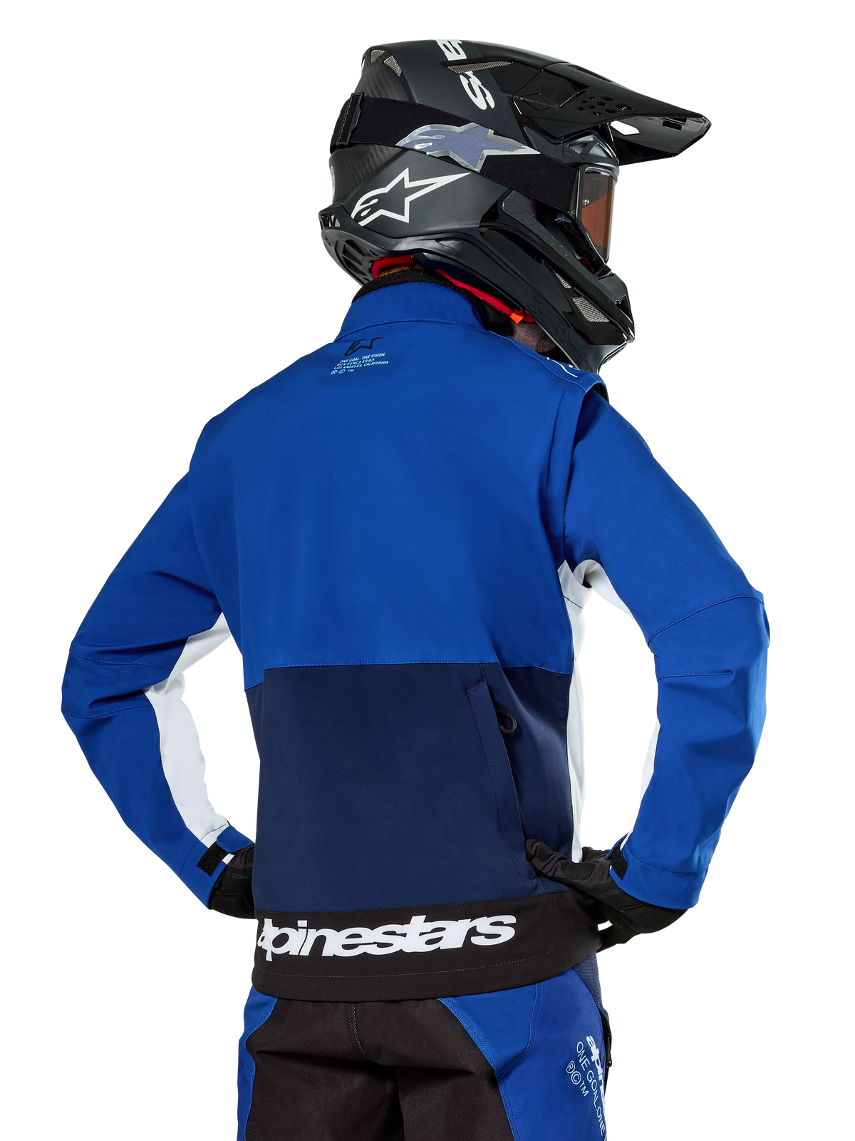 Giacca Softshell Alpinestars Lite-Dura in blu navy, azzurro e bianco, vista posteriore che mostra la grande tasca posteriore con zip per riporre le maniche, inserti laterali bianchi e marchio Alpinestars sull'orlo, indossata con casco da motocross nero e attrezzatura coordinata.