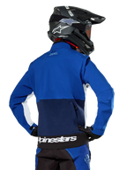 Giacca Softshell Alpinestars Lite-Dura in blu navy, azzurro e bianco, vista posteriore che mostra la grande tasca posteriore con zip per riporre le maniche, inserti laterali bianchi e marchio Alpinestars sull'orlo, indossata con casco da motocross nero e attrezzatura coordinata.