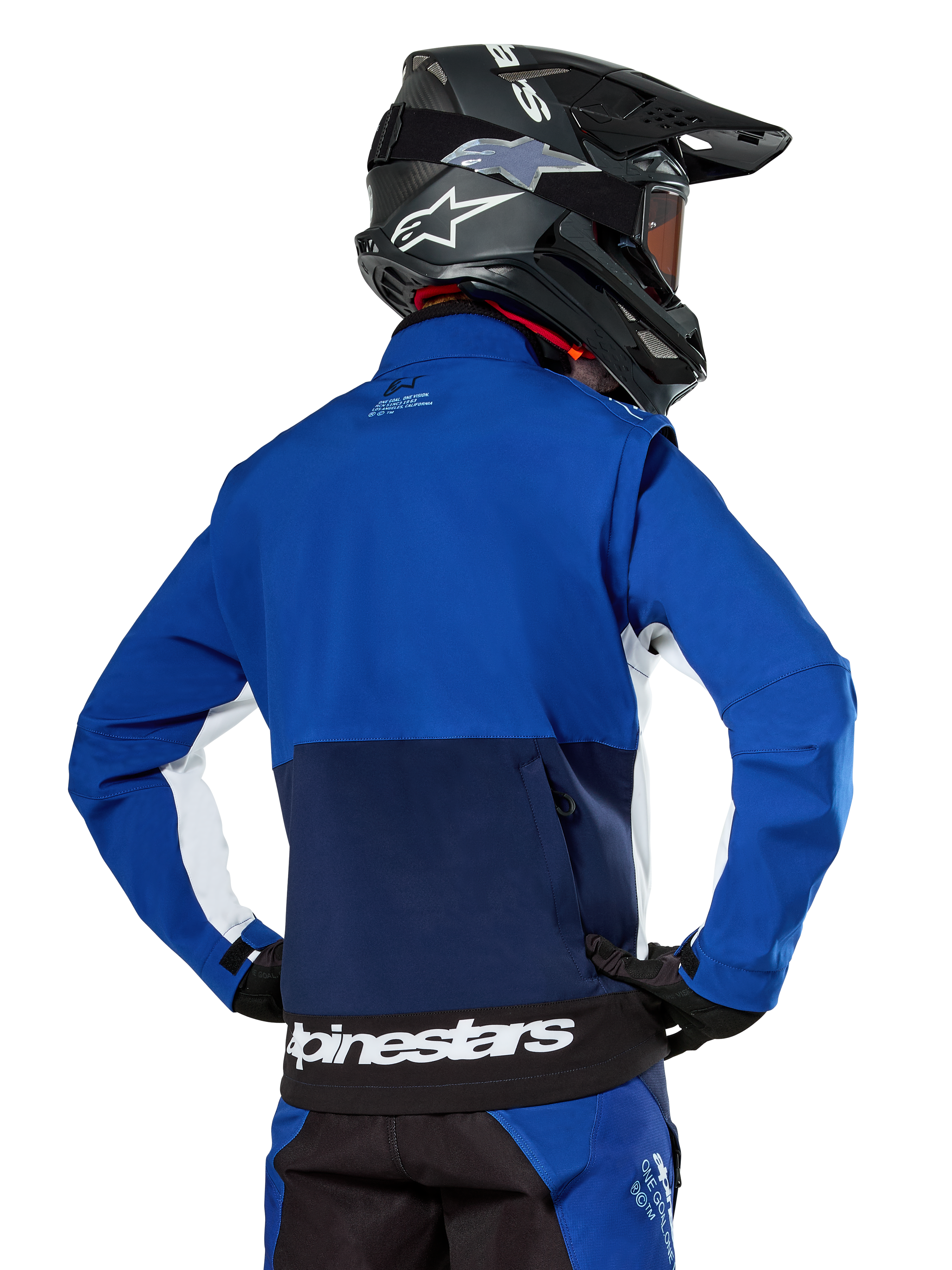 Giacca Softshell Alpinestars Lite-Dura in blu navy, azzurro e bianco, vista posteriore che mostra la grande tasca posteriore con zip per riporre le maniche, inserti laterali bianchi e marchio Alpinestars sull'orlo, indossata con casco da motocross nero e attrezzatura coordinata.