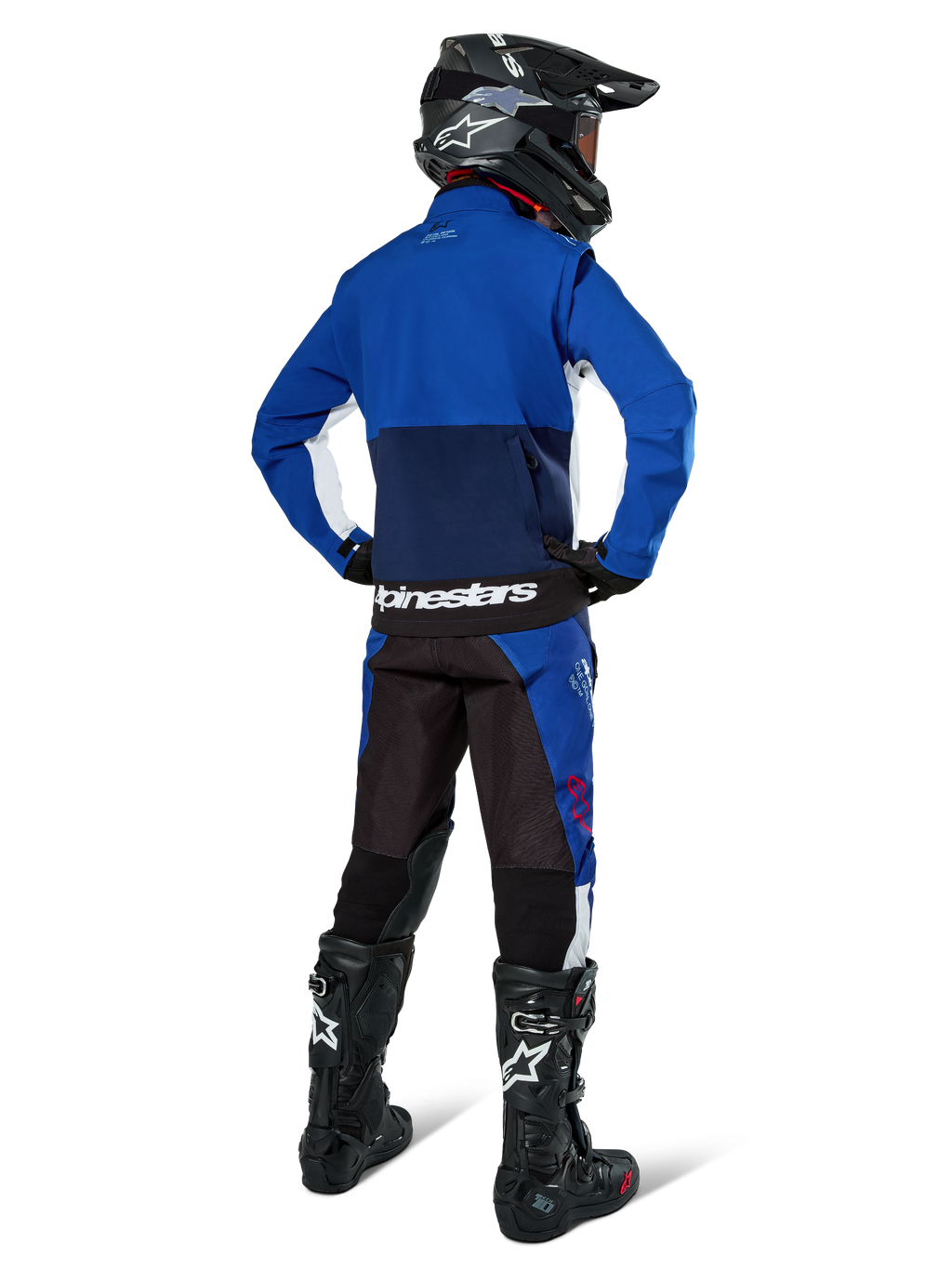 Pilota di motocross visto di spalle che indossa la giacca Alpinestars Lite-Dura Softshell in Navy Bright Blue e bianco, abbinata a pantaloni da enduro coordinati, casco nero e stivali neri, abbigliamento completo da gara off-road