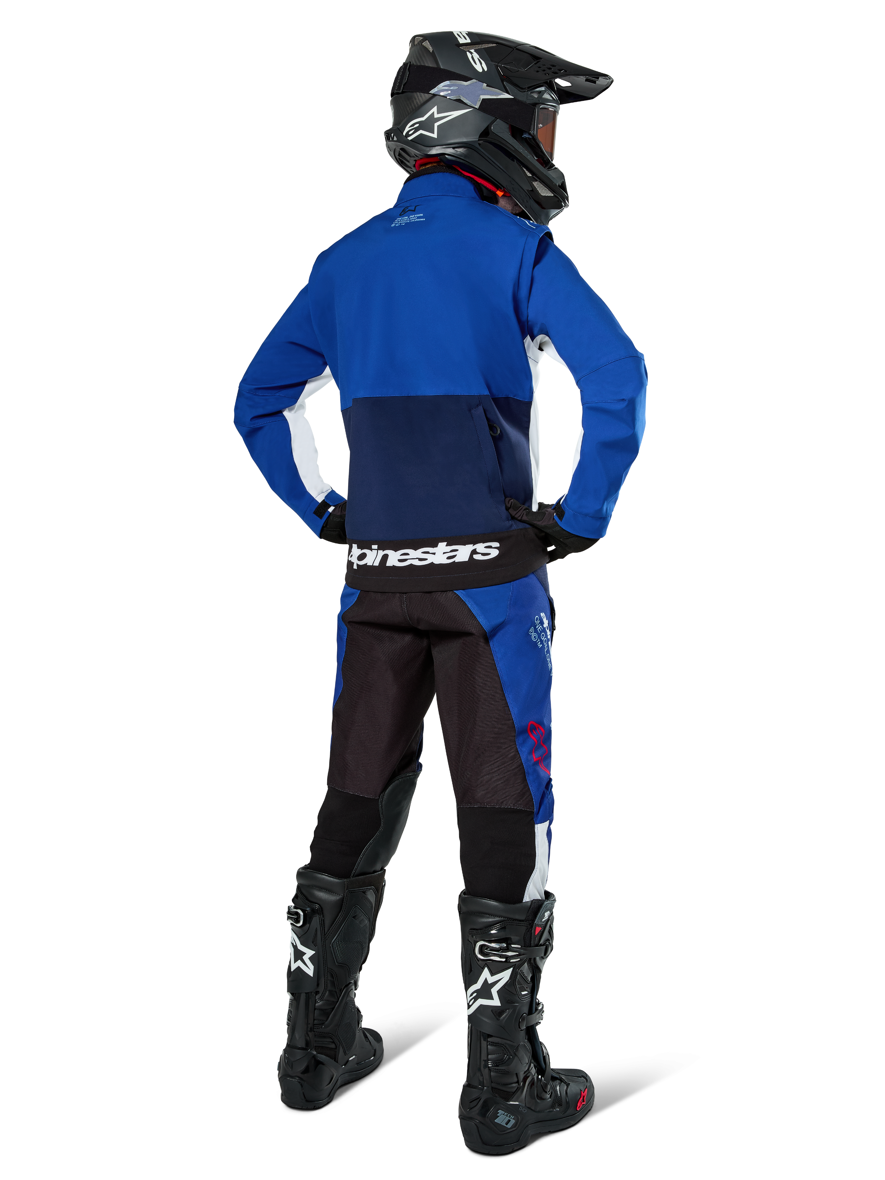 Pilota di motocross visto di spalle che indossa la giacca Alpinestars Lite-Dura Softshell in Navy Bright Blue e bianco, abbinata a pantaloni da enduro coordinati, casco nero e stivali neri, abbigliamento completo da gara off-road