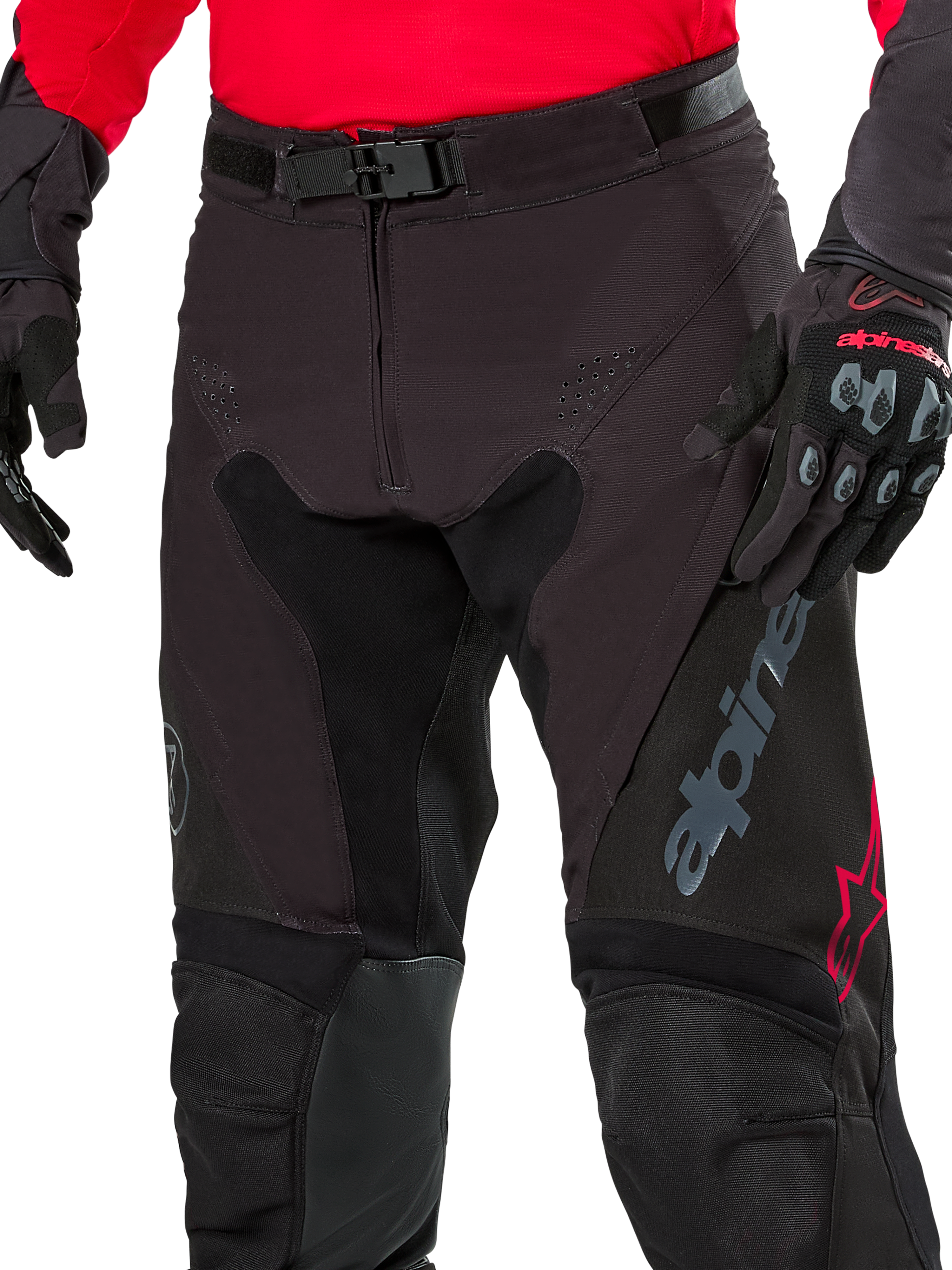 Primo piano dei pantaloni Alpinestars Pro-Dura, pantaloni da enduro, nero con accenti rossi, dotati di chiusura sicura con fibbia magnetica e patta con cerniera, indossati con maglia rossa coordinata e guanti neri, robusta costruzione in ripstop per guida off-road