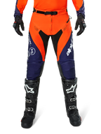 Pantaloni Alpinestars Pro-Dura, pantaloni da motocross ed enduro, blu navy, arancione acceso e bianco, con pannelli neri e loghi bianchi, indossati con stivali e guanti da fuoristrada neri, abbigliamento protettivo professionale per trail e off-road