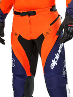 Alpinestars Pro-Dura Pants, pantaloni da moto enduro, blu navy, arancione acceso e bianco, vista del dettaglio della vita con fibbia magnetica SX2 e prese d'aria con zip sulle cosce, materiale ripstop resistente con branding evidente, indossati con abbigliamento coordinato