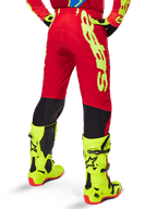 Alpinestars 2025 Supertech Maker Pants, pantaloni da motocross, rosso acceso e giallo fluo, vista posteriore che mostra il grande logo giallo sul lato e i pannelli neri sulla parte inferiore delle gambe, indossati con stivali e attrezzatura giallo fluo coordinati