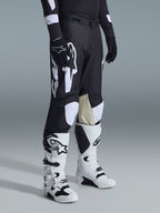Pantaloni Alpinestars Racer Air Riway 2026, pantaloni da motocross neri e bianchi con grandi grafiche del logo bianco, pannelli interni delle ginocchia in pelle beige, indossati con stivali off-road bianchi e neri e guanti neri in studio.