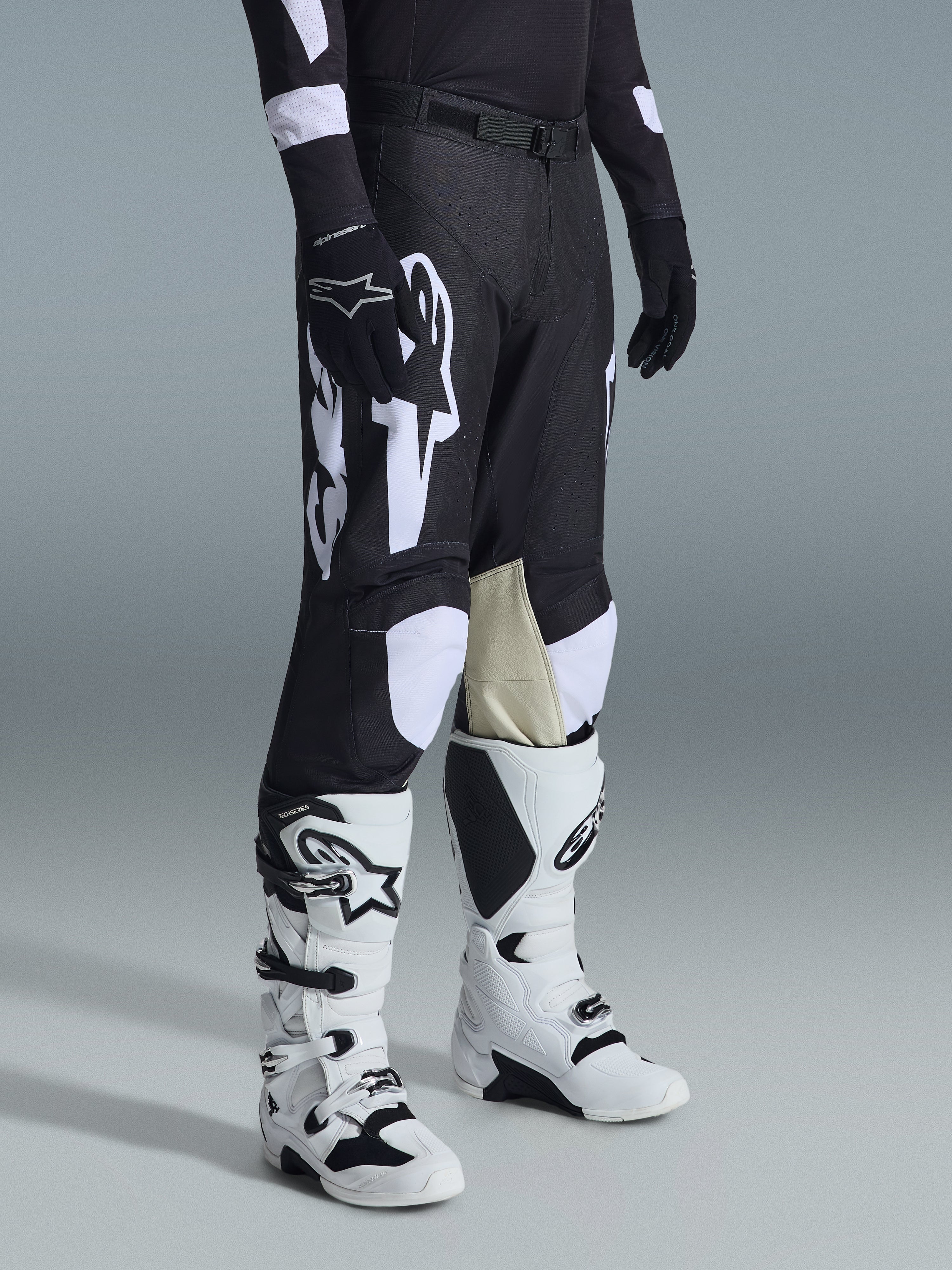 Pantaloni Alpinestars Racer Air Riway 2026, pantaloni da motocross neri e bianchi con grandi grafiche del logo bianco, pannelli interni delle ginocchia in pelle beige, indossati con stivali off-road bianchi e neri e guanti neri in studio.