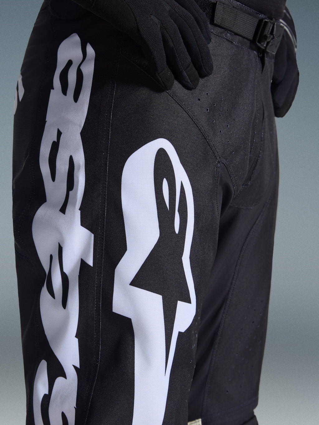 Pantaloni Alpinestars Racer Air Riway 2026, pantaloni da motocross neri con grande logo Alpinestars bianco sulla coscia, primo piano del tessuto tecnico e della chiusura regolabile in vita, indossati con guanti neri per la guida fuoristrada